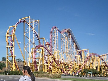 Estructura de una montaña rusa de color amarillo y vía morada ante un cielo azul con una persona en primer plano