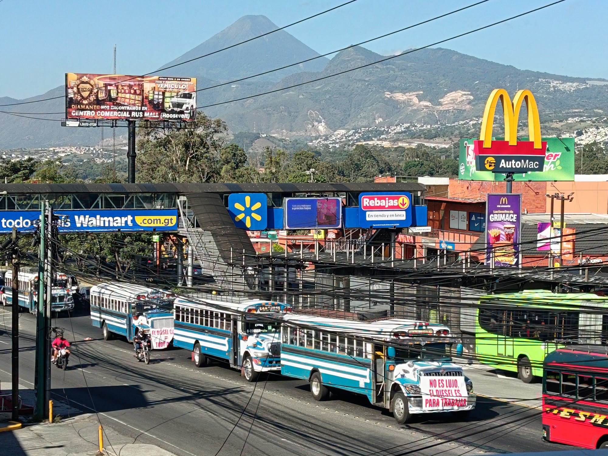 Varias unidades de transporte público transitan por una calzada en Guatemala con pancartas sobre el costo del diésel. (Redes Sociales)
