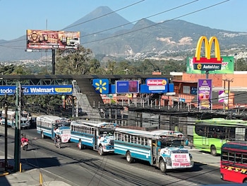 Autobuses azules y blancos circulan por una carretera con vallas publicitarias, cables de electricidad y montañas al fondo