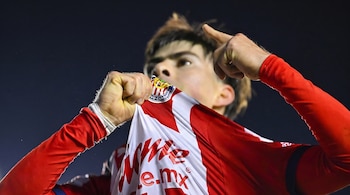 Primer plano de un futbolista con camiseta rojiblanca de Chivas que levanta el cuello y señala con el dedo índice hacia arriba.