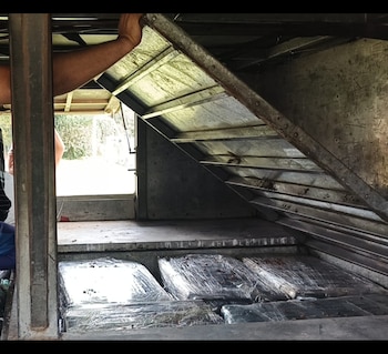 Varios paquetes rectangulares envueltos en plástico transparente yacen en un compartimento oculto bajo una estructura metálica; se ve el brazo de una persona