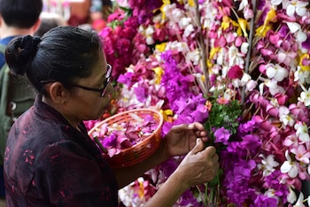 Mujer con gafas y cabello recogido, en ropa oscura, sostiene cesta con pétalos. Arregla un mural de flores artificiales fucsia, moradas, blancas y amarillas