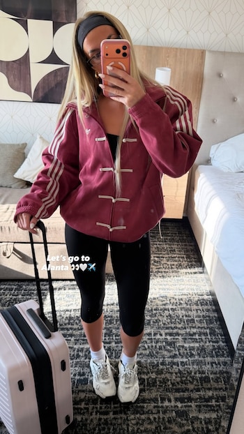 Mujer rubia con chaqueta deportiva roja, leggings negros y zapatillas blancas se toma una selfie en un espejo, sostiene una maleta rosa y un teléfono