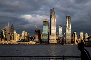 Horizonte de Nueva York con rascacielos brillando bajo un cielo oscuro y nublado. Río en primer plano con una barandilla y la silueta de una persona a la derecha