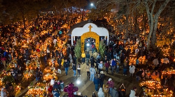 Vista aérea nocturna de personas en un cementerio con tumbas iluminadas por velas, flores amarillas, árboles y una pequeña capilla central.