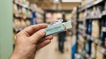 Una mano sostiene un chicle verde con envoltorio plateado abierto en el pasillo de un supermercado con estantes al fondo.