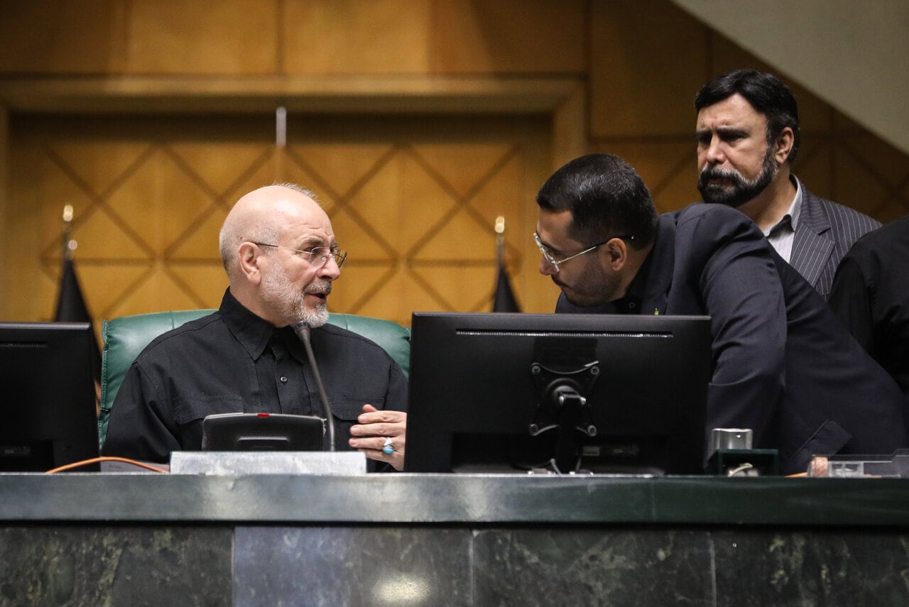 Mohammad Bagher Ghalibaf speaks with a man leaning across the presiding desk while another official stands behind them at the Islamic Consultative Assembly in Tehran, Iran, on October 25, 2026. Speaker Mohammad Bagher Ghalibaf chaired the October 25 session, which combined physical attendance at the Islamic Consultative Assembly with participation by video link.