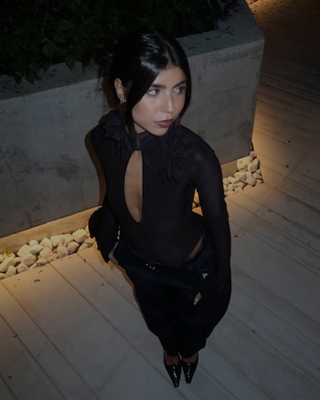 Cami Mayan sonríe sosteniendo un ramo de flores envuelto en papel blanco, vestida con top oscuro, jeans y cinturón dorado, frente a unas escaleras de madera