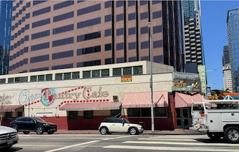 Fachada de un edificio de una planta con el letrero Original Pantry Cafe, toldos a rayas, rascacielos al fondo y automóviles en la calle