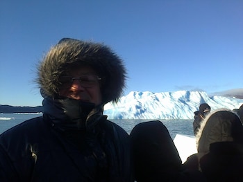 Hombre con abrigo de invierno y capucha de piel, gafas, en un entorno de agua, con un gran iceberg y montañas al fondo, bajo cielo azul