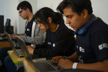 Tres personas sentadas en fila utilizan computadoras portátiles sobre mesas individuales en una sala