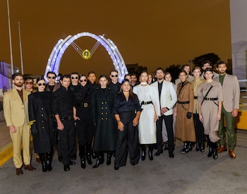 Grupo de veintiún personas, entre hombres y mujeres, posa con trajes y abrigos en tonos neutros; al fondo, un arco blanco iluminado de noche