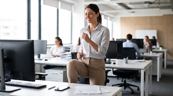 Mujer en blusa y pantalón beige, levanta la rodilla derecha en una oficina con ventanas grandes y monitores de computadora. Hay personas borrosas de fondo.