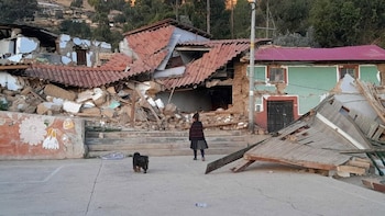 Una niña de espaldas y un perro en una explanada de cemento frente a dos edificios parcialmente derrumbados y un tercero dañado