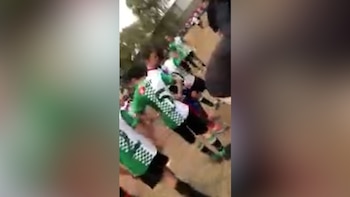 Varias personas con uniformes de cuadros verdes y blancos y pantalones negros se alinean en un entorno exterior de tierra con árboles desenfocados