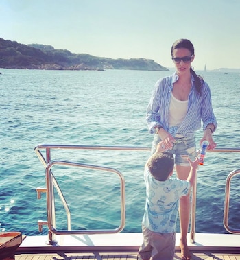 Una mujer con gafas de sol y camisa de rayas junto a un niño pequeño en la cubierta de un barco. Detrás se ve el mar azul y una costa montañosa