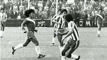 Fotografía en blanco y negro de Diego Maradona y varios futbolistas en una cancha de fútbol, con un balón en el aire y público detrás