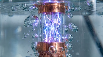 Primer plano de un tubo de vidrio recubierto de cobre en un líquido, con brillantes descargas eléctricas azules y moradas visibles dentro del tubo y burbujas alrededor.