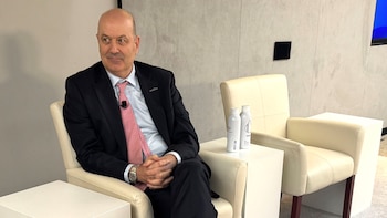 Un hombre calvo con traje oscuro y corbata rosa está sentado en un sillón crema, con las manos juntas y mirando hacia la izquierda