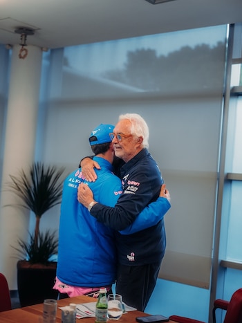 Un hombre mayor de cabello blanco y gafas abraza a un hombre joven con gorra azul y chaqueta del equipo Alpine, en un interior con mesa