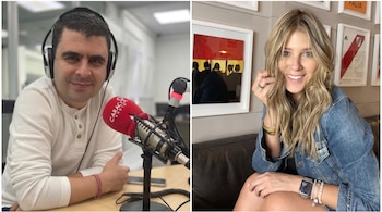 Mujer rubia con cabello ondulado y largo sonríe mirando al frente, lleva chaqueta de jean, reloj inteligente y anillos. Al fondo hay cuadros decorativos y una camiseta de fútbol.
