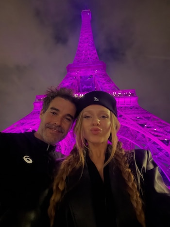 Un hombre y una mujer se toman una foto en primer plano con la Torre Eiffel iluminada de morado en el fondo