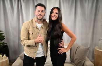 Nicolas Magaldi, con chaqueta beige, y Pampita, con vestido negro, posan sonrientes con los brazos alrededor. Se ven cortinas grises y un sofá claro