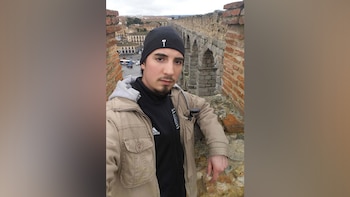 Un hombre con gorro negro y chaqueta beige posa sobre un muro de ladrillos. Detrás, un acueducto de piedra con arcos y edificios de ciudad