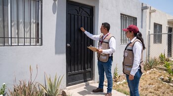 Dos trabajadores, un hombre y una mujer, con chalecos beige y credenciales oficiales, tocan la puerta de una casa gris con rejas. El hombre sujeta un portapapeles y una tableta.