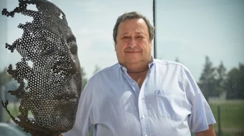 Un hombre sonriente con camisa a rayas azul claro se sitúa junto a una escultura metálica de un rostro fragmentado, con un fondo exterior borroso