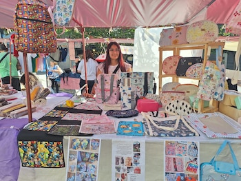 Mujer joven sonríe detrás de un puesto de mercado con una carpa rosa. El puesto está lleno de bolsos, mochilas y monederos coloridos con diseños variados