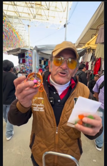 Hombre de mediana edad con gorra, gafas de sol amarillas y chaleco, sostiene un juguete colorido y una bolsa en un mercado al aire libre Foto: eltripon1 (Captura de pantalla Instagram)