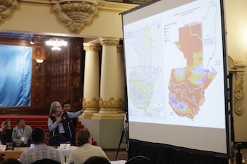 Una mujer habla por micrófono y señala una pantalla donde proyectan dos mapas de Guatemala ante un público sentado en mesas