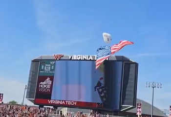 Un paracaidista con una bandera de Estados Unidos descendiendo frente a una pantalla gigante de Virginia Tech en un estadio, bajo un cielo azul
