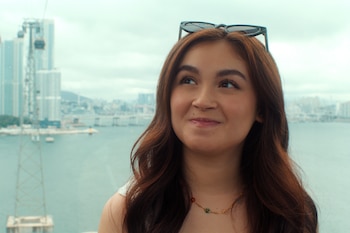 Primer plano de Anna Cathcart con gafas de sol sobre su cabello castaño, mirando hacia arriba. Al fondo se ven edificios borrosos, agua y un teleférico