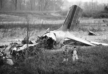 Restos de la cola de un avión destrozado en un campo. Dos hombres caminan en primer plano, otros operarios trabajan cerca de los restos