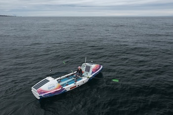 Persona en bote de remo blanco y colorido, con paneles solares, remando en el mar. Cielo nublado y línea de costa distante
