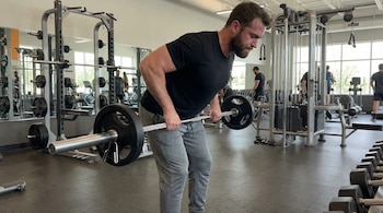 Hombre musculoso de unos 30 años, sudoroso, inclinado hacia adelante sosteniendo una barra con pesas con agarre en pronación en un gimnasio.