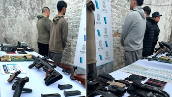 Hombres de espaldas junto a una mesa con armas de fuego, municiones, teléfonos, dinero en efectivo y un banner de la Policía