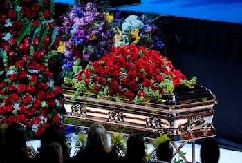 Ataúd dorado con rosas rojas y follaje verde en la tapa, frente a grandes arreglos florales. Varias personas se observan en primer plano