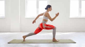 Mujer con top y leggings claros en colchoneta verde, haciendo ejercicio de zancada. Sus muslos y glúteos muestran una representación anatómica en rojo