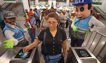 Mujer pasando tarjeta por torniquete de acceso, acompañada de personal con chaleco azul de ATU y una mascota de ATU, en una estación de transporte