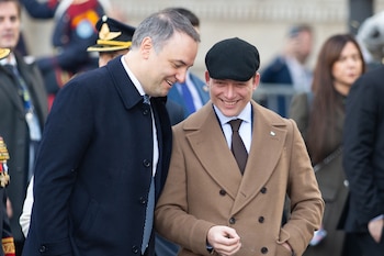 Primer plano de dos hombres blancos sonriendo y mirando hacia abajo a la derecha; uno lleva un abrigo azul oscuro, el otro un abrigo marrón y boina negra