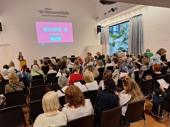 Interior de un auditorio con una ponente en un podio y una audiencia sentada frente a una pantalla que muestra 'Voluntariado +60'