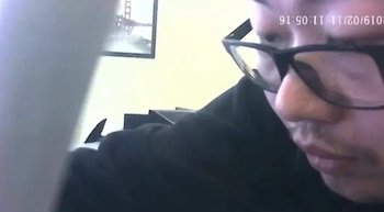 Un hombre con gafas y ropa negra mira hacia abajo frente a una cámara que registra una marca de tiempo en la parte superior
