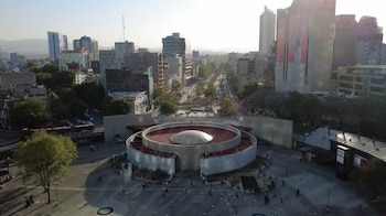 Vista aérea de una plaza circular con una estructura abovedada en el centro, rodeada de avenidas, árboles, vehículos y edificios urbanos