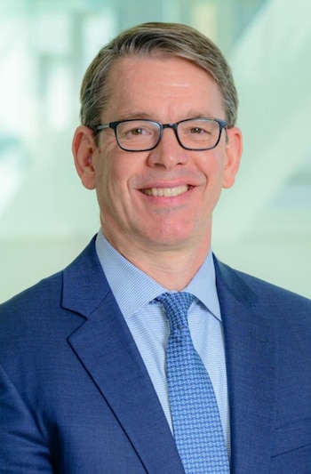 Retrato de un hombre sonriente con gafas, saco de vestir azul, camisa clara y corbata azul