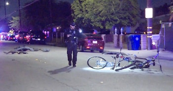Un policía camina de noche por una calle donde hay dos bicicletas caídas en el asfalto, restos de vehículos y patrullas con luces al fondo