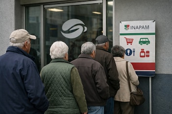 Cuatro adultos mayores de espaldas, con cabello canoso y ropa abrigada, forman una fila frente a la puerta de vidrio de un banco y un cartel de INAPAM.