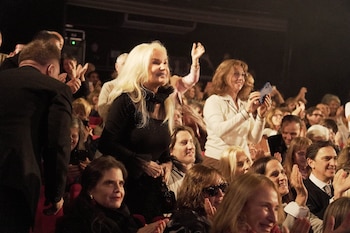 Susana Giménez, con cabello rubio largo y blusa negra, sonríe y aplaude en un teatro. Varias personas a su alrededor también aplauden en la audiencia